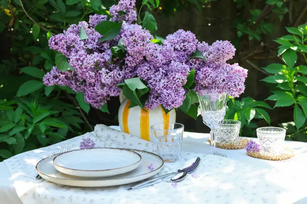 Bahçedeki masada güzel leylak çiçekleri, tabaklar, çatal bıçaklar, bardaklar ve mumlar