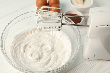 Krem şantili kase, el mikseri ve malzemeler beyaz ahşap masada, yakın plan.