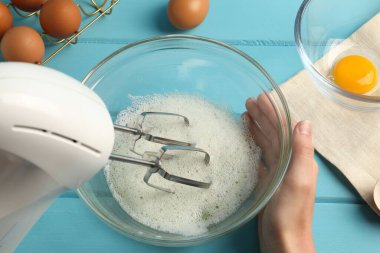Kadın, açık mavi ahşap masada el çırpıcı ile yumurta beyazı çırpıyor.