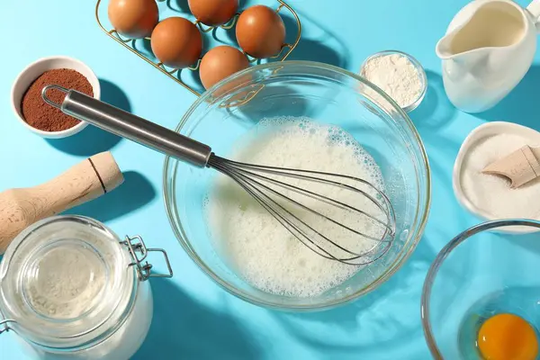 Fırça çırpma, yumurta beyazı kasede ve diğer ürünler açık mavi masada, düz yerde.