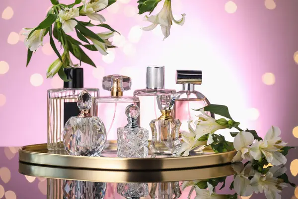Ayna yüzeyinde birçok farklı parfüm şişesi ve çiçek var. Pembe arka plan, bulanık ışıklar, yakın plan.