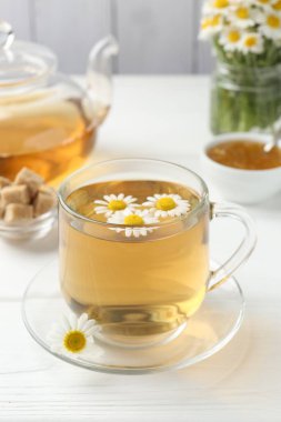 Cam fincanda nefis papatya çayı, kahverengi şeker, çaydanlık, reçel ve taze çiçekler beyaz ahşap masada, yakın plan.