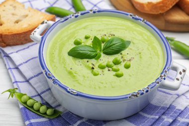 Beyaz ahşap masada servis edilen fesleğenli bezelye kremalı çorba.
