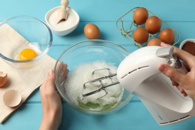 Bir kadın açık mavi ahşap masada el çırpıcı ile yumurta beyazı çırpıyor.