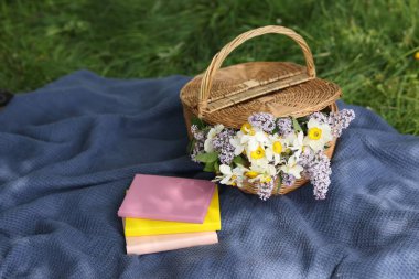 Yığınla kitap, battaniye ve hasır sepet. Yeşil çimlerin üzerinde çiçekler.