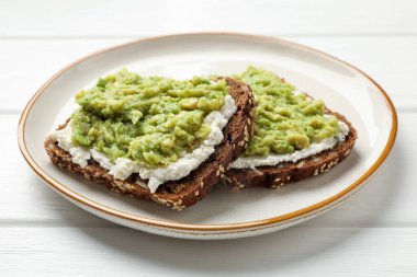 Avokado ve krem peynirli lezzetli sandviçler beyaz ahşap masada, yakın plan.