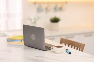 Laptop, kırtasiye malzemeleri ve bir fincan sıcak içecek mutfaktaki beyaz mermer masada, seçici bir odak noktası