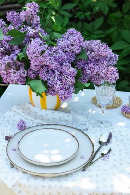 Bahçedeki masada güzel leylak çiçekleri, tabaklar, çatal bıçaklar ve bardaklar.