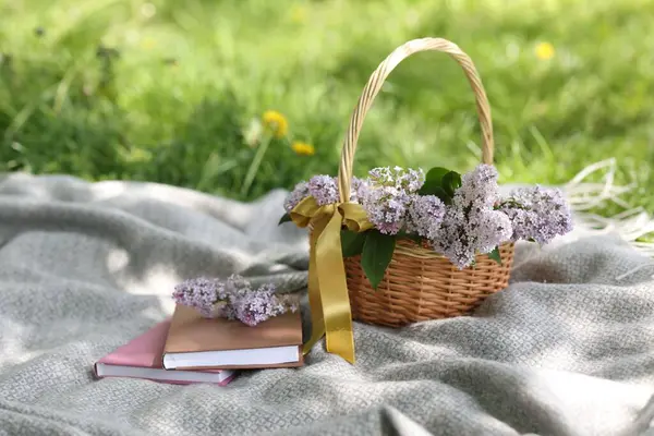 Yığınla kitap ve hasır sepet. Yeşil çimlerin üzerinde leylak çiçekleri.