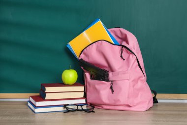 Sırt çantasında silah, kırtasiye malzemeleri, bardaklar ve elma tahtanın yanındaki tahta masada.