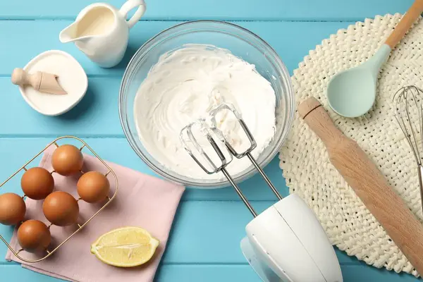 Açık mavi ahşap masa üzerinde çırpılmış krema, yumurta ve diğer ürünlerle el çırpıcı düz yatıyordu.