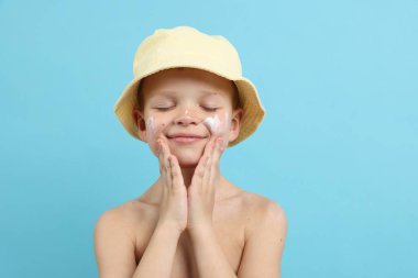 Küçük çocuk açık mavi arka planda cildine güneş kremi sürüyor.