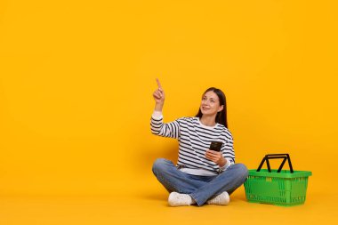 Akıllı telefonlu ve alışveriş sepetli gülümseyen kadın sarı arka plandaki bir şeyi işaret ediyor. Metin için boşluk
