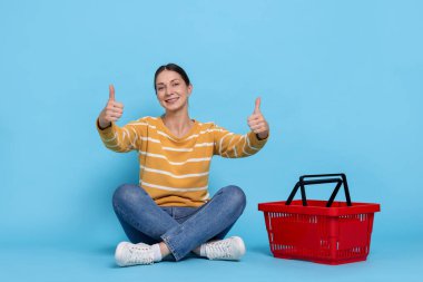 Gülümseyen kadın başparmağını kaldırıyor ve açık mavi arka planda alışveriş sepeti alıyor.