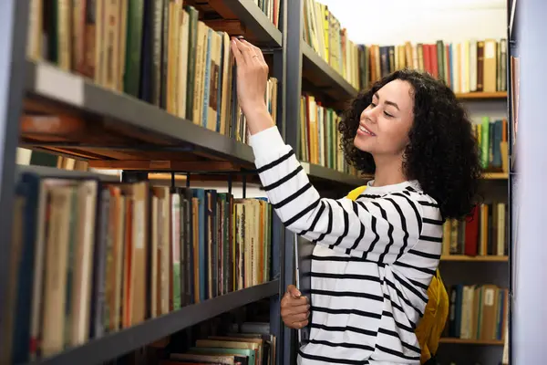 Bilgisayarlı bir kadın kütüphanedeki raftan kitap seçiyor.