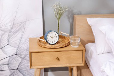 Komodinin yanında çalar saat, vazoda çiçekler ve yatağın yanında bir bardak su.