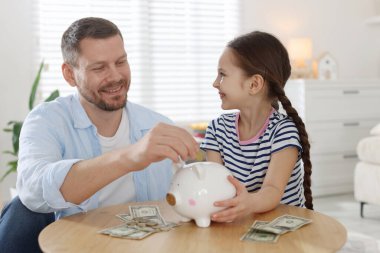 Baba ve kızı kapalı bir masadaki kumbaraya cep harçlığı koyuyor.