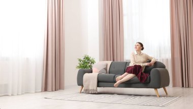 Kadın pencerenin yanındaki kanepede oturuyor ve evinde şık perdeler var. Metin için boşluk