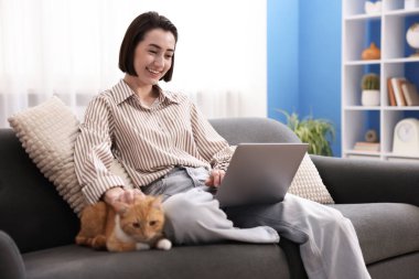 Bilgisayarla çalışan gülümseyen kadın ve ev ofisindeki kanepede kediyi okşayan kadın.