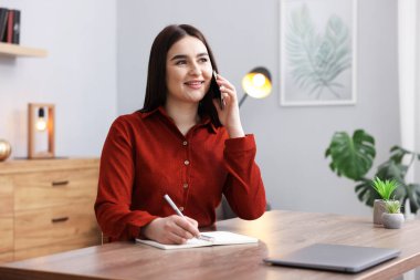 Planlayıcılı gülümseyen kadın notlar alıyor ve ahşap masada akıllı telefondan konuşuyor.