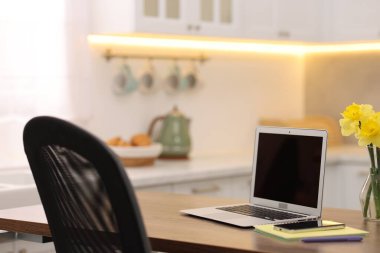 Boş ekranlı dizüstü bilgisayar, akıllı telefon, kırtasiye malzemeleri ve mutfaktaki ahşap masada çiçekler. Tasarım için model