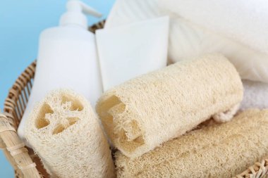 Açık mavi arka planda farklı lif süngerleri, kozmetik ürünleri ve havlular sepette.