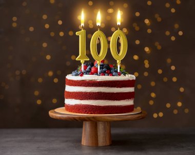 Böğürtlenli ve yanan mumlarla kaplı nefis bir pasta. Arka planda koyu renkli ve bulanık ışıklar var. 100. yaş günü veya başka özel bir gün.