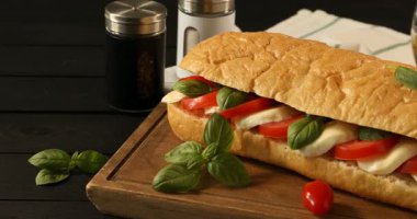 Mozarella peynirli lezzetli sandviç, domates ve fesleğen siyah ahşap masada, kamera sağa hareket ediyor.