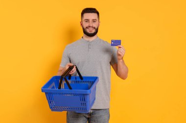 Boş alışveriş sepeti ve turuncu arka planda kredi kartı olan bir adam.