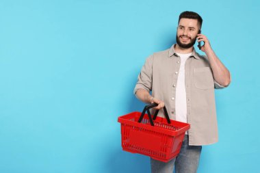 Boş alışveriş sepetli bir adam akıllı telefondan açık mavi arkaplana, mesaj için yere karşı konuşuyor.