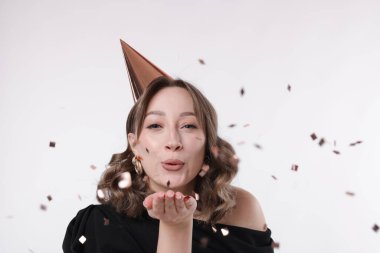 Doğum günü kutlaması. Parti konisi olan bir kadın beyaz arka planda konfeti üflüyor.