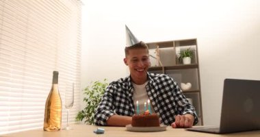 Doğum günü kutlaması. Adam pastaya mum üflerken içerideki masada dizüstü bilgisayarla görüntülü arama yapıyor.