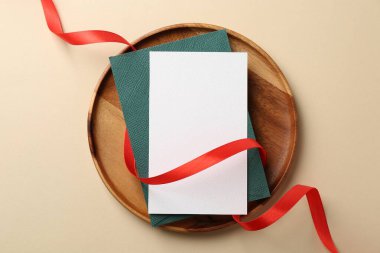 Boş davetiye kartı, zarf, kırmızı kurdele ve bej arka planda ahşap tepsi.