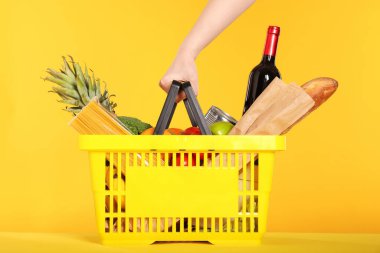 Sarı arka planda ürünlerle dolu alışveriş sepeti olan bir kadın.