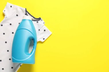 Bir şişe yumuşatıcı ve sarı arka planda bebek kıyafetleri. Metin için boşluk