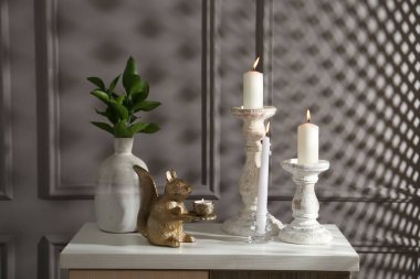 Zarif şamdanlar, yanan mumlar, vazoda dallar ve gri duvarın yanındaki şifoniyerde diğer dekorlar.