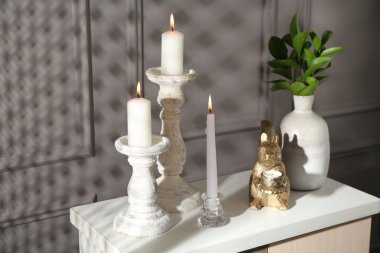 Zarif şamdanlar, yanan mumlar, vazoda dallar ve gri duvarın yanındaki şifoniyerde diğer dekorlar.