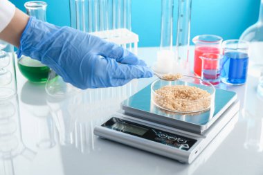 Petri kabını tartan bilim adamı beyaz ayna masasındaki numunelerle açık mavi arkaplanı karşılaştırıyor.