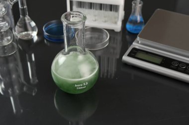 Yeşil sıvılı matara, diğer laboratuvar camları ve ölçekli siyah ayna masası, yakın plan.