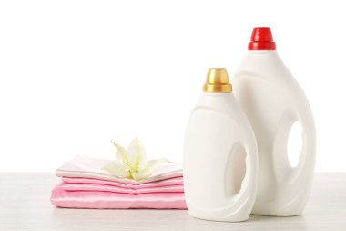 Kumaş yumuşatıcı şişeler, üst üste dizilmiş elbiseler ve çiçek beyaz arka plana karşı ahşap masa