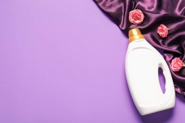 Bir şişe yumuşatıcı, çiçekler ve mor arka planda renkli kumaş, düz yatak. Metin için boşluk