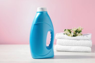 Bir şişe yumuşatıcı, terry havlular ve beyaz ahşap masada pembe arka planda zambak çiçekleri.