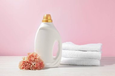 Bir şişe yumuşatıcı, havlular ve karanfil çiçekleri beyaz ahşap masa üzerinde pembe arka planda.