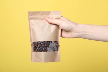 Sarı arka planda kahve çekirdekli fermuarlı stand-up çantası tutan kadın, yakın plan. Tasarım için model