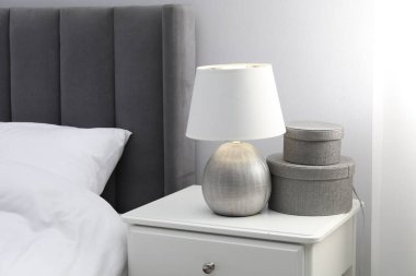 Yatağın yanında lamba ve dekor olan bir komodin.