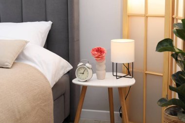 Yan masada lamba, çalar saat ve yatağın yanında çiçek var.