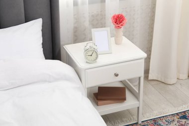 Yatağın yanında çalar saati, çiçeği ve çerçevesi olan bir komodin.