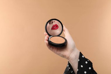 Kırmızı dudaklı güzel kadın pudra aynasına bakıyor koyu bej arka planda, yakın plan