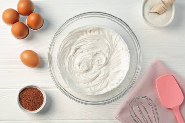 Krem şantili kase, çırpılmış çırpılmış ve malzemeler beyaz ahşap masada, düz yatıyordu.