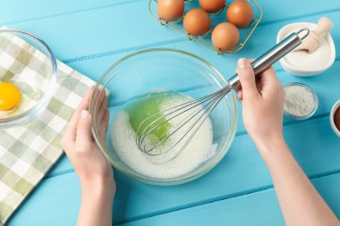 Kadın açık mavi ahşap masada kasede yumurta beyazı çırpıyor.